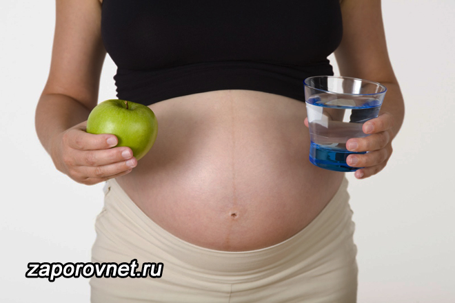 Беременная девушка держит в одной руке стакан воды, в другой - яблоко
